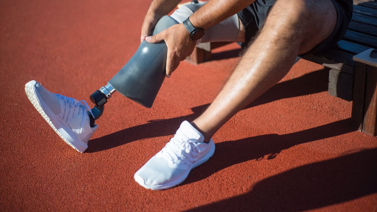 An adult man sitting on a bench adjusting his prosthetic leg on an outdoor track.
