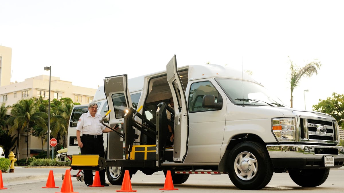 A driver standing by a wheelchair accessible van with open doors and a ramp, surrounded by cones.