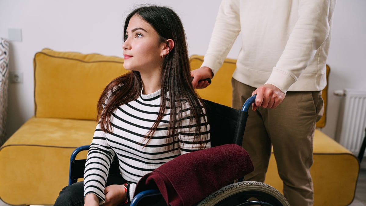 A young woman in a wheelchair being assisted indoors by a caregiver, showcasing support and mobility.